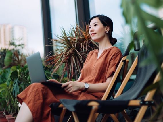 Contemplated young Asian woman looking away in thought while relaxing on deck chair using laptop in the backyard, surrounded by beautiful houseplants. Lifestyle and technology