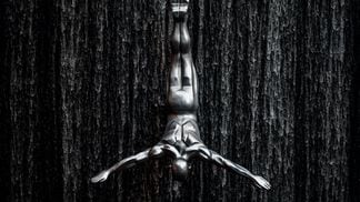 Black and white photo of a sculpture diving/hanging in front of water.