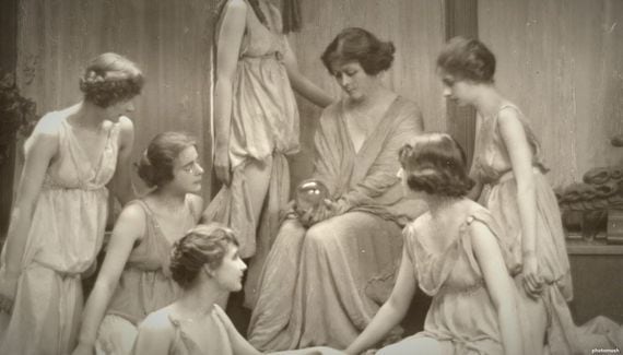 Group portrait centered on Isadora Duncan, seated with her hands in her lap holding a crystal ball and gazing downwards. She is surrounded by six other female dancers who are posing in standing, kneeling, or sitting positions, and all gazing at her. In the background are four curtained windows and potted plants.