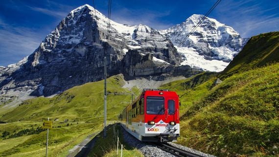 A train travels through the Swiss Alps with snowy peaks in the background.